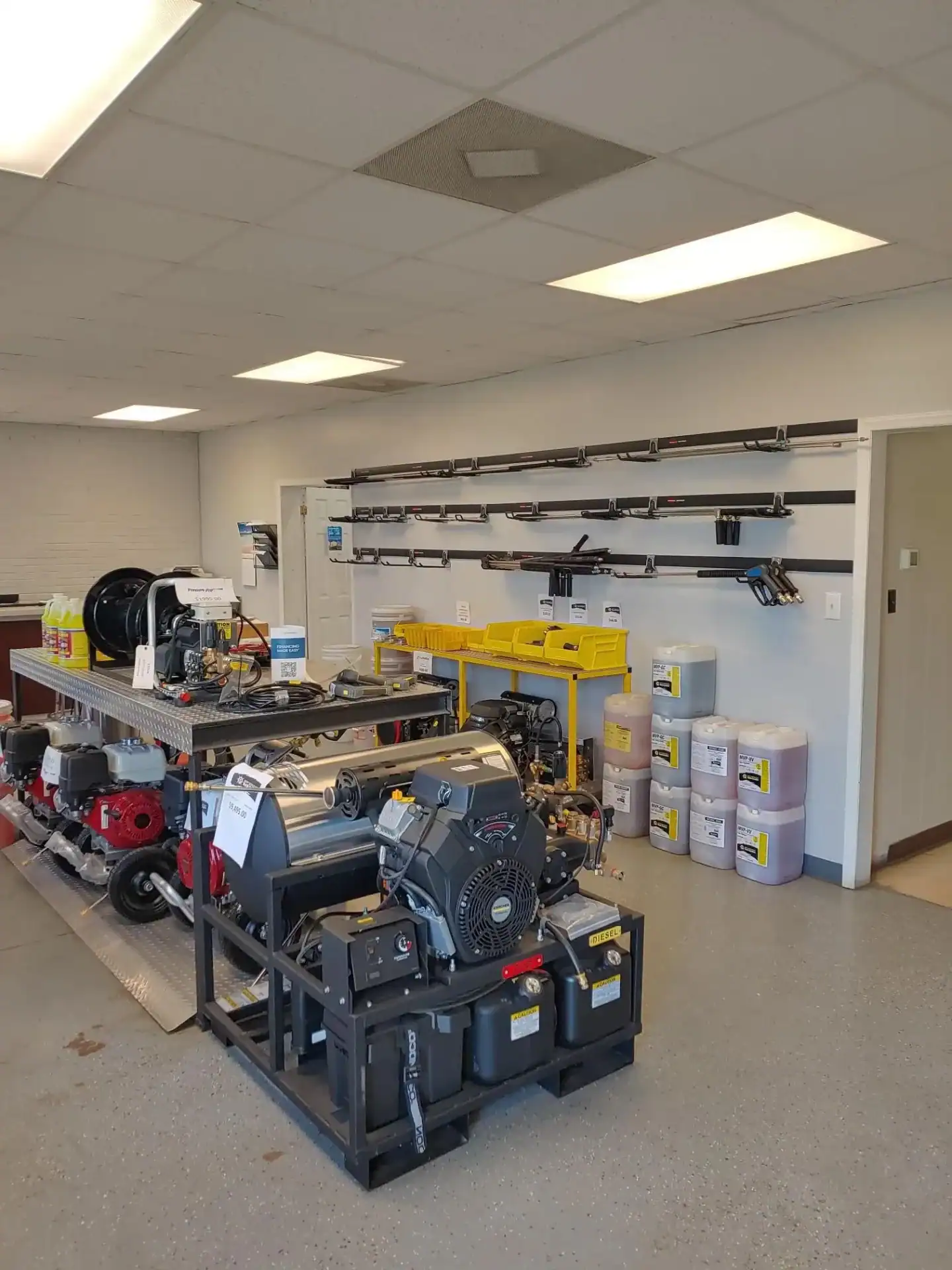 A well-lit workshop with power equipment, cleaning machines, chemical containers, and tools organized on wall racks and shelves.