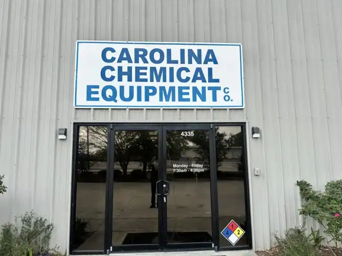 Entrance to Carolina Chemical Equipment Co. building with a large sign above glass doors and business hours posted; hazard placard is visible near the door.