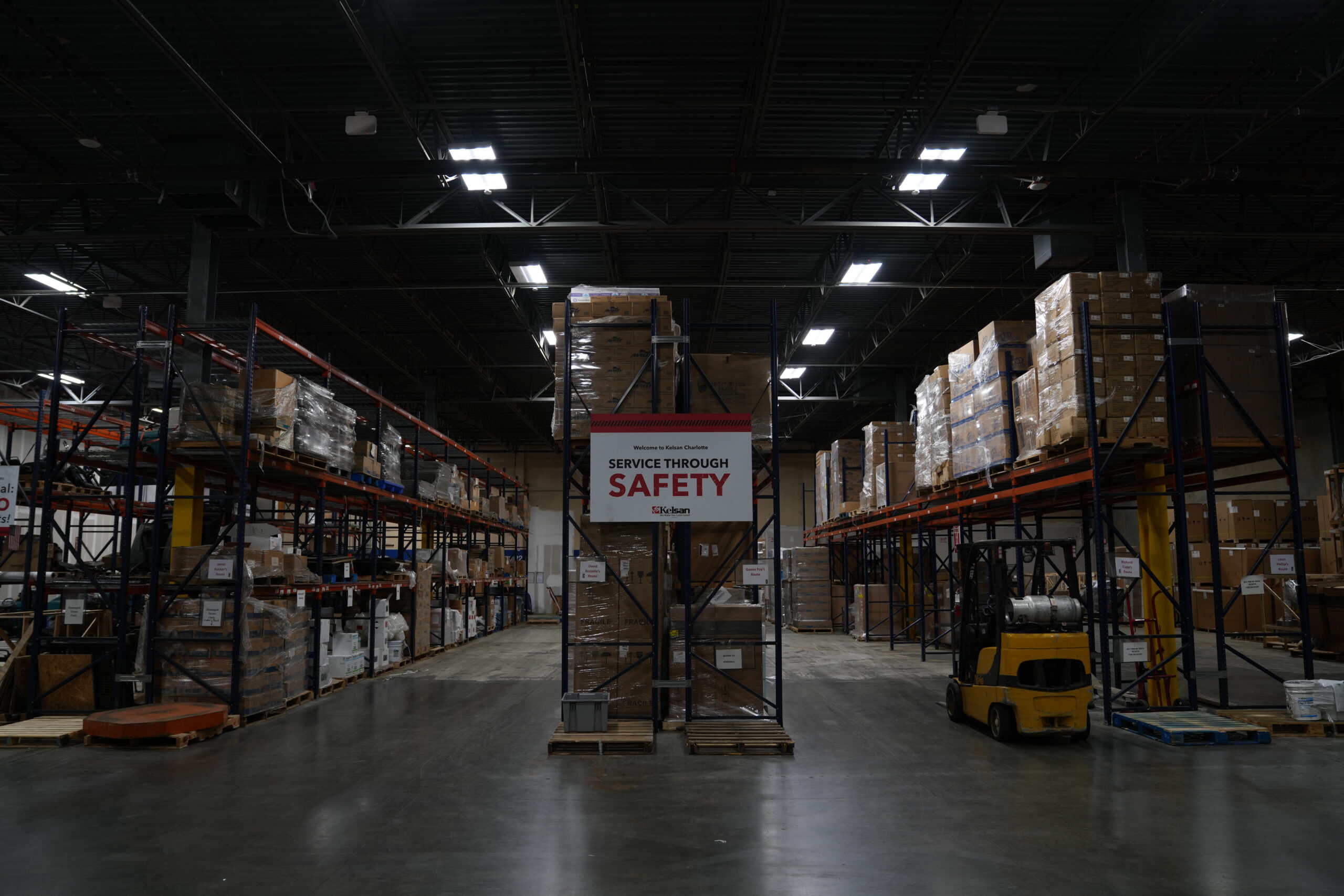 A warehouse with shelves stacked with boxes, a forklift, and a sign reading "Service Through Safety" in the center aisle.
