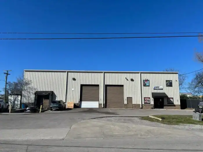 A beige industrial warehouse with two large garage doors, a small entrance door, parked cars, and a clear blue sky overhead.
