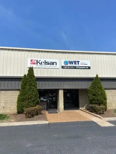 Exterior view of a commercial building with two signs above the entrance for Kelsan and WET Equipment, flanked by two tall evergreen shrubs.