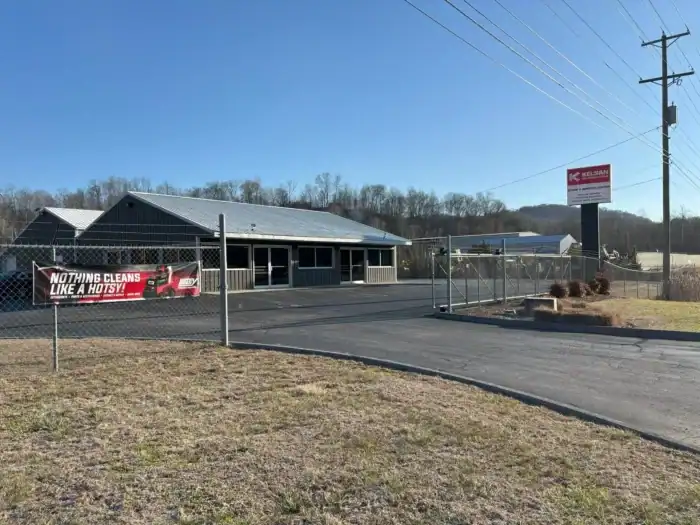 A fenced commercial building with a banner reading "NOTHING CLEANS LIKE A HOTSY!!" and a sign labeled "KELSAN" stands beside a paved driveway on a clear day.