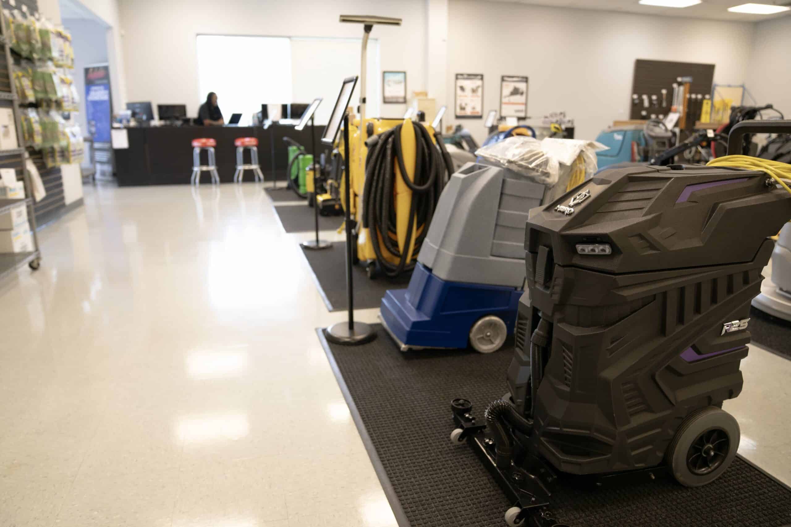 A showroom displays various commercial cleaning machines on mats; a person sits at a desk in the background near a bright window.