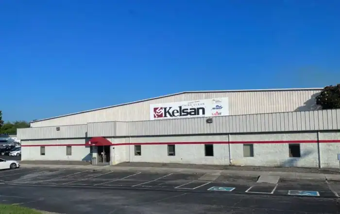 A large industrial building with a "Kelsan" sign on the front, an empty parking lot, and a clear blue sky overhead.