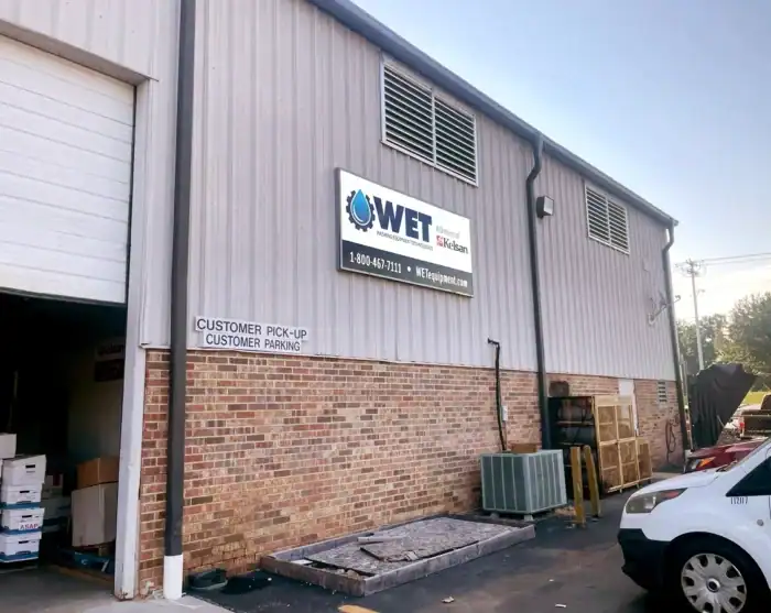 Exterior of a commercial building with a large WET Equipment Solutions sign, a customer parking sign, and a partially open garage door with boxes visible inside.