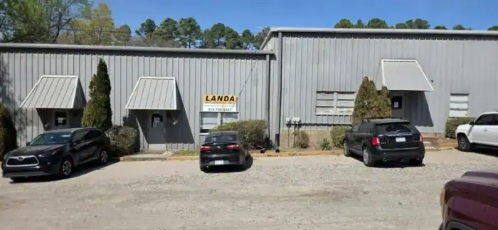 A small gray metal building with a "LANDA" sign above the entrance, three parked cars, and some shrubs in front.