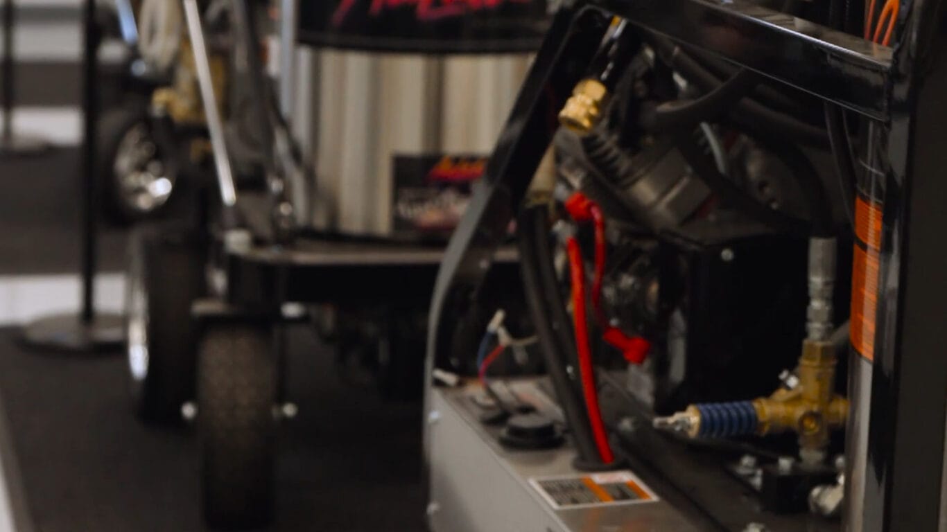 Close-up of mechanical equipment with visible wires, hoses, and metal components; a wheeled machine is blurred in the background.